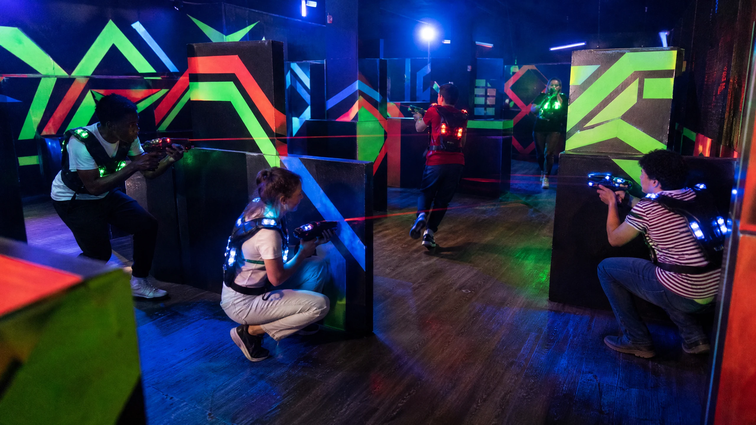 Spieler gehen beim Lasertag hinter Hindernissen in Deckung und zielen mit Laserpistolen durch einen neonbunten Raum