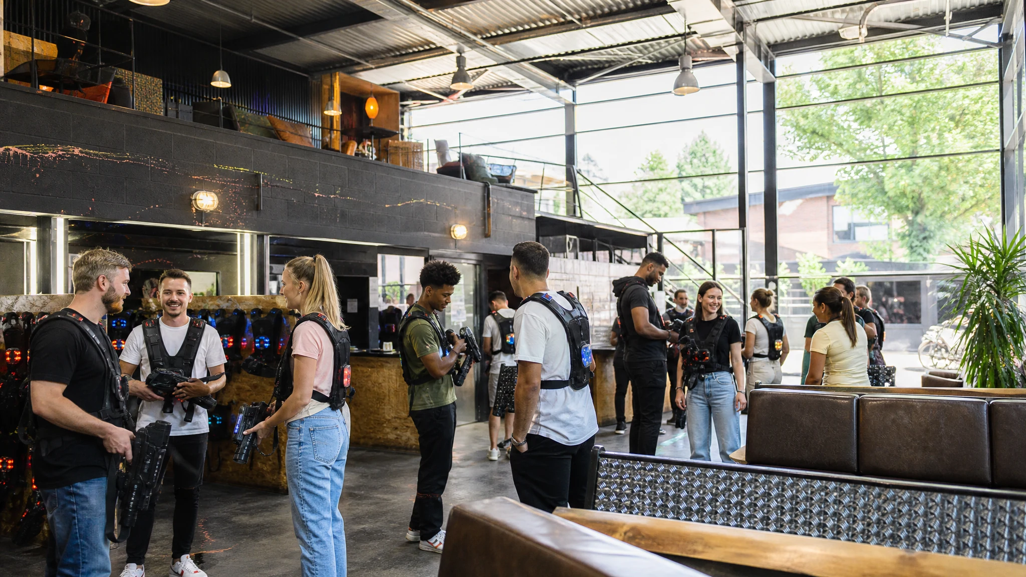 Gäste mit Westen und Spielgeräten warten vor einer Runde Lasertag in einer hellen Halle mit Sofas und Glasfront