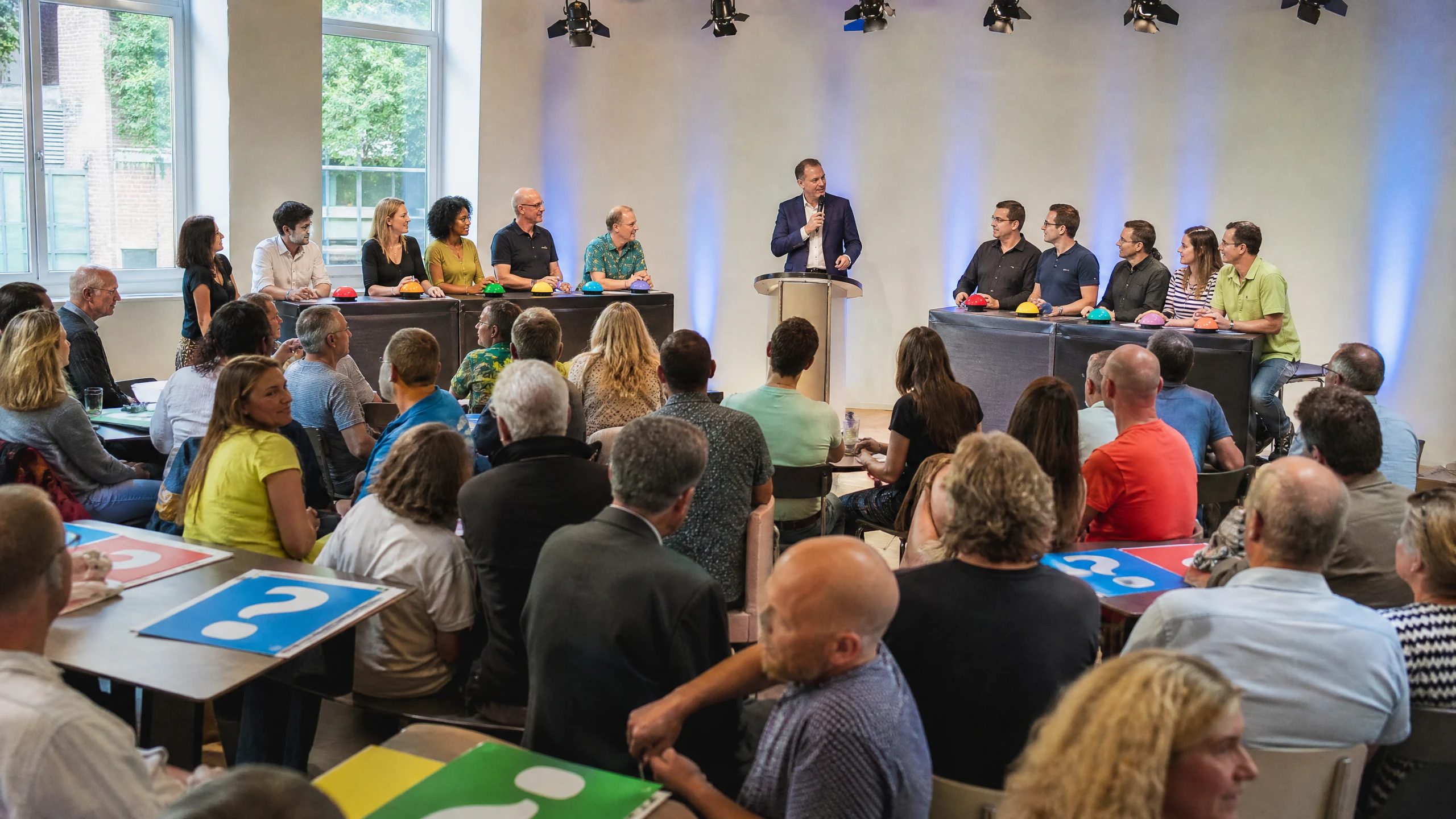 Moderator mit Mikrofon vor Rategruppen an Pulten mit Buzzern, Publikum im Saal bei einer Quiz Game Show