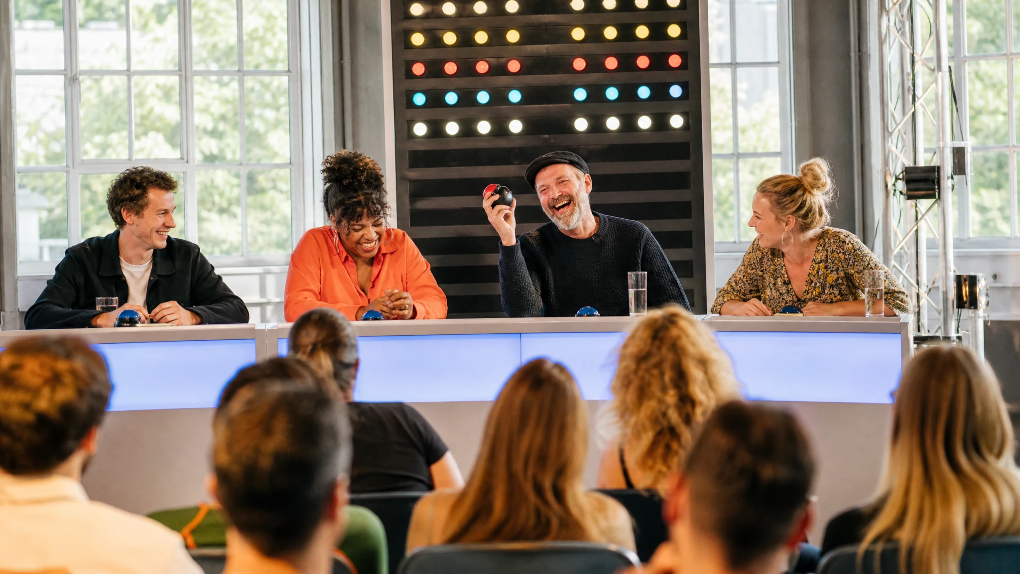 Lachende Rategäste mit Buzzern an einem Pult vor Publikum in einem Studio, wie bei Quiz Game Shows mit Comedy