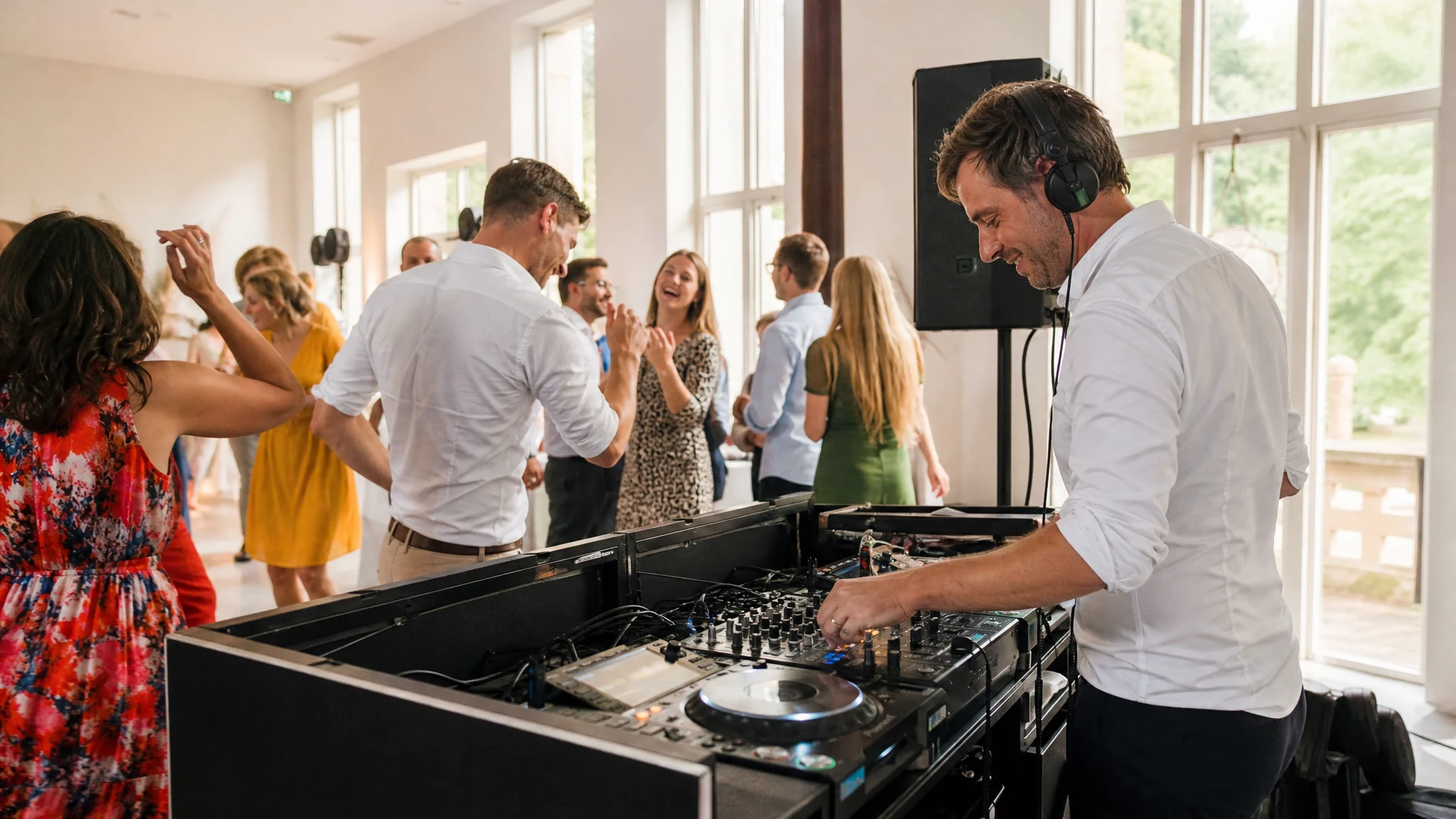 Ein DJ mit Kopfhörern lächelt am Mischpult, während Gäste in einem hellen Saal auf einer Hochzeit in Bonn tanzen