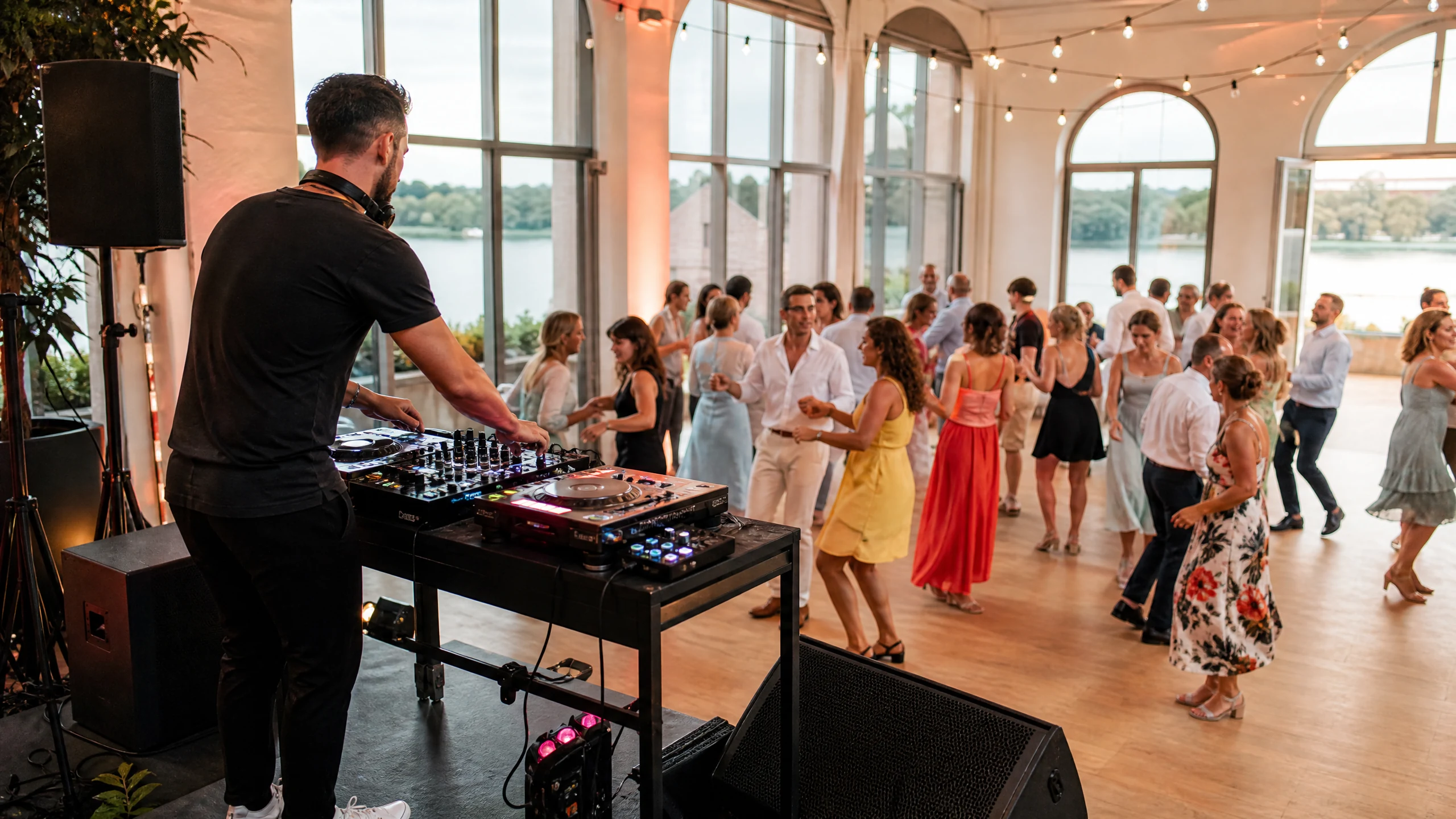 Ein DJ legt bei einer Hochzeit in Bonn am Mischpult auf, während Gäste in festlicher Kleidung in einem Saal am Wasser tanzen
