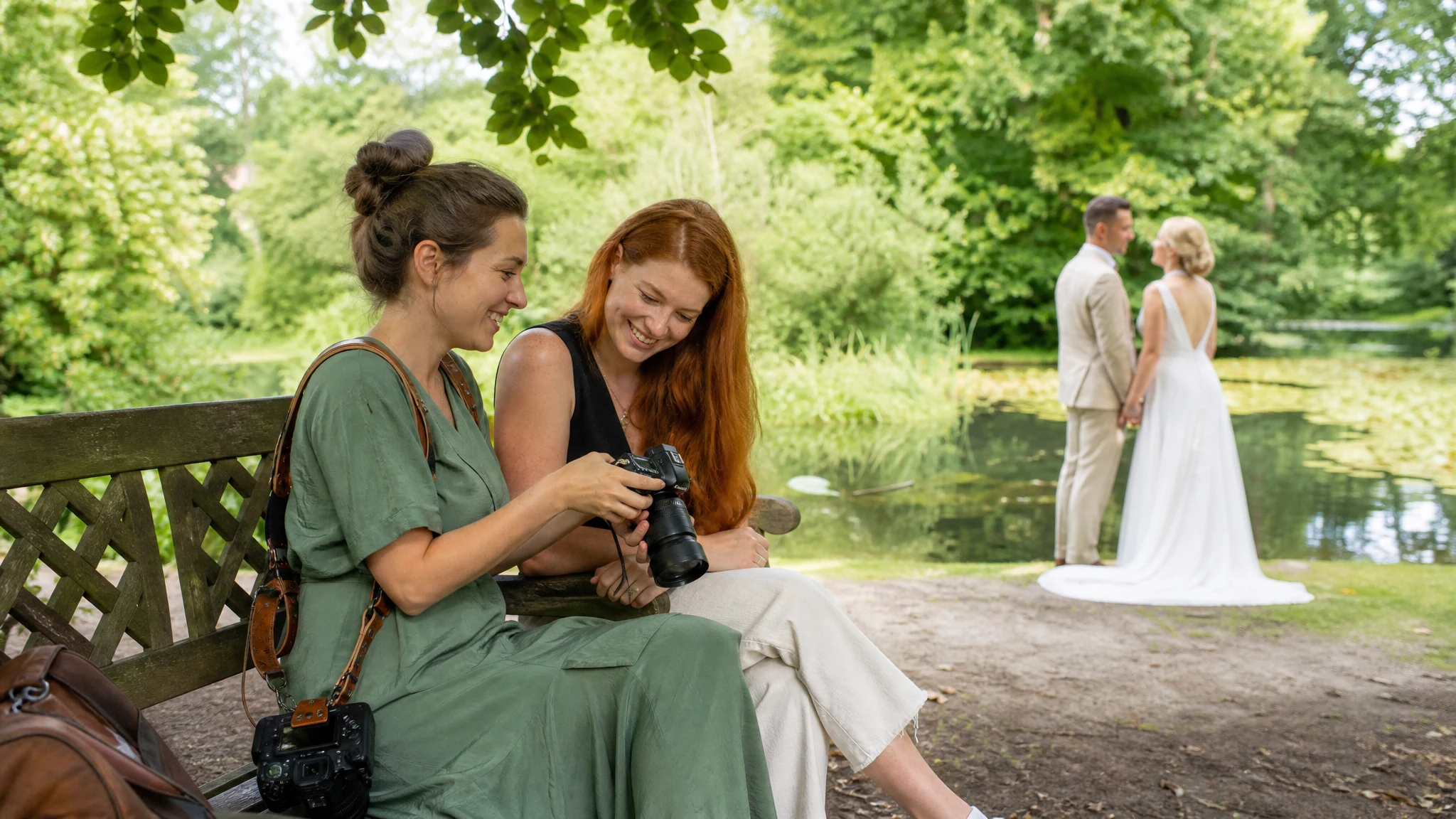 Eine Hochzeitsfotografin in Bonn zeigt einer Kollegin lachend Bilder auf der Kamera, im Park steht ein Brautpaar am Teich