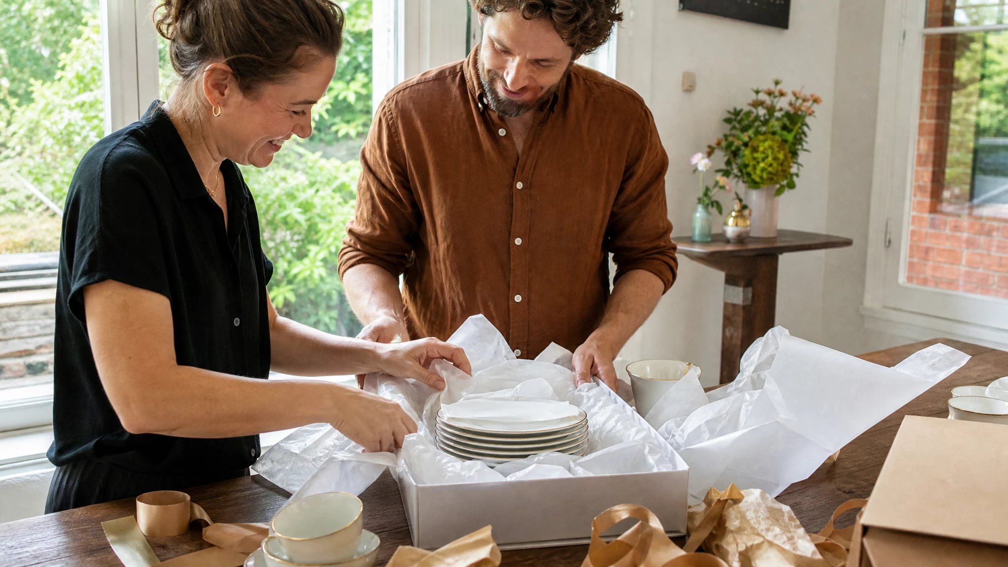 Eine Frau und ein Mann packen lachend Porzellanteller als Geschenk zum 20. Hochzeitstag aus Seidenpapier aus