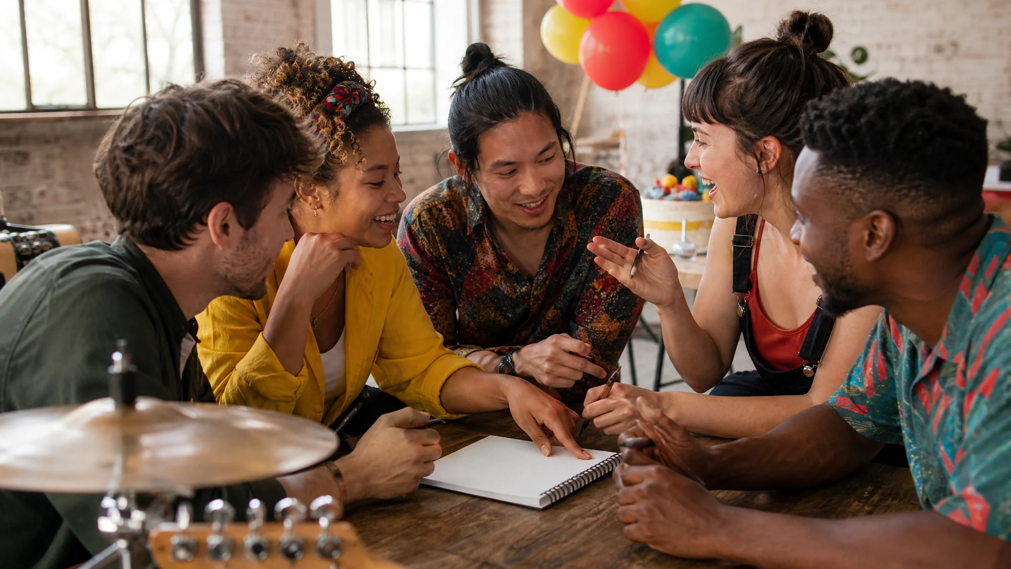 Freunde planen lachend am Holztisch mit Notizbuch eine Band für einen Geburtstag, dahinter hängen Luftballons