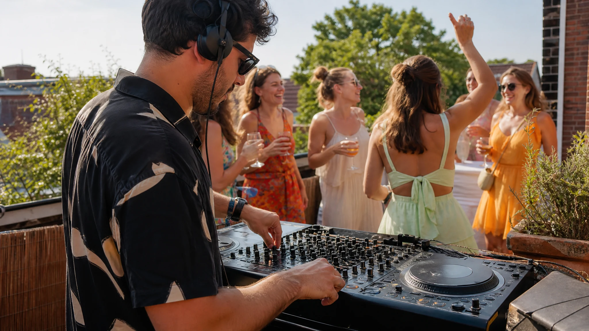 Ein DJ mit Kopfhörern legt auf einer Dachterrasse beim JGA auf, Gäste mit Getränken tanzen und lachen im Hintergrund