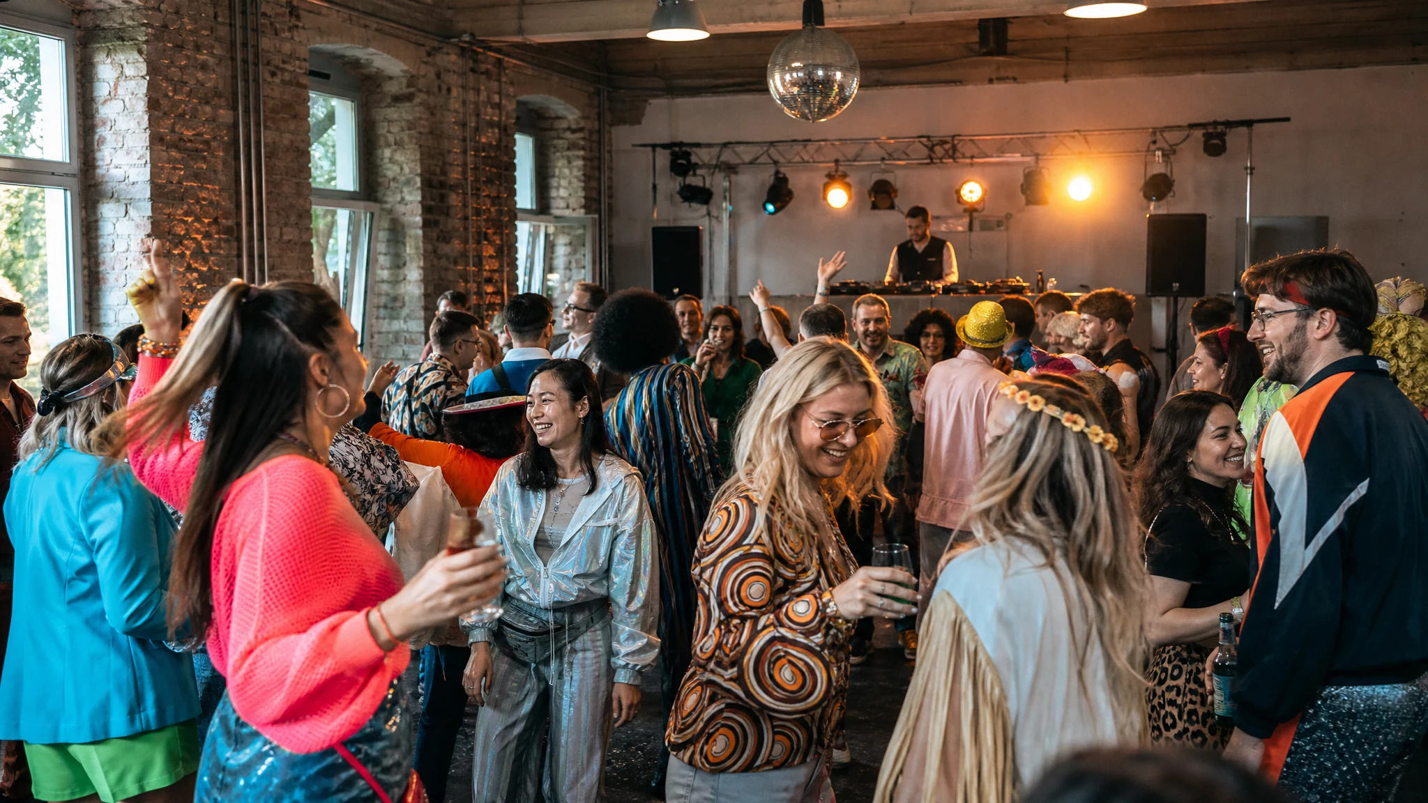 Gäste in bunten Retro-Outfits tanzen und lachen in einer Backsteinhalle, ein DJ legt auf einer Mottoparty auf