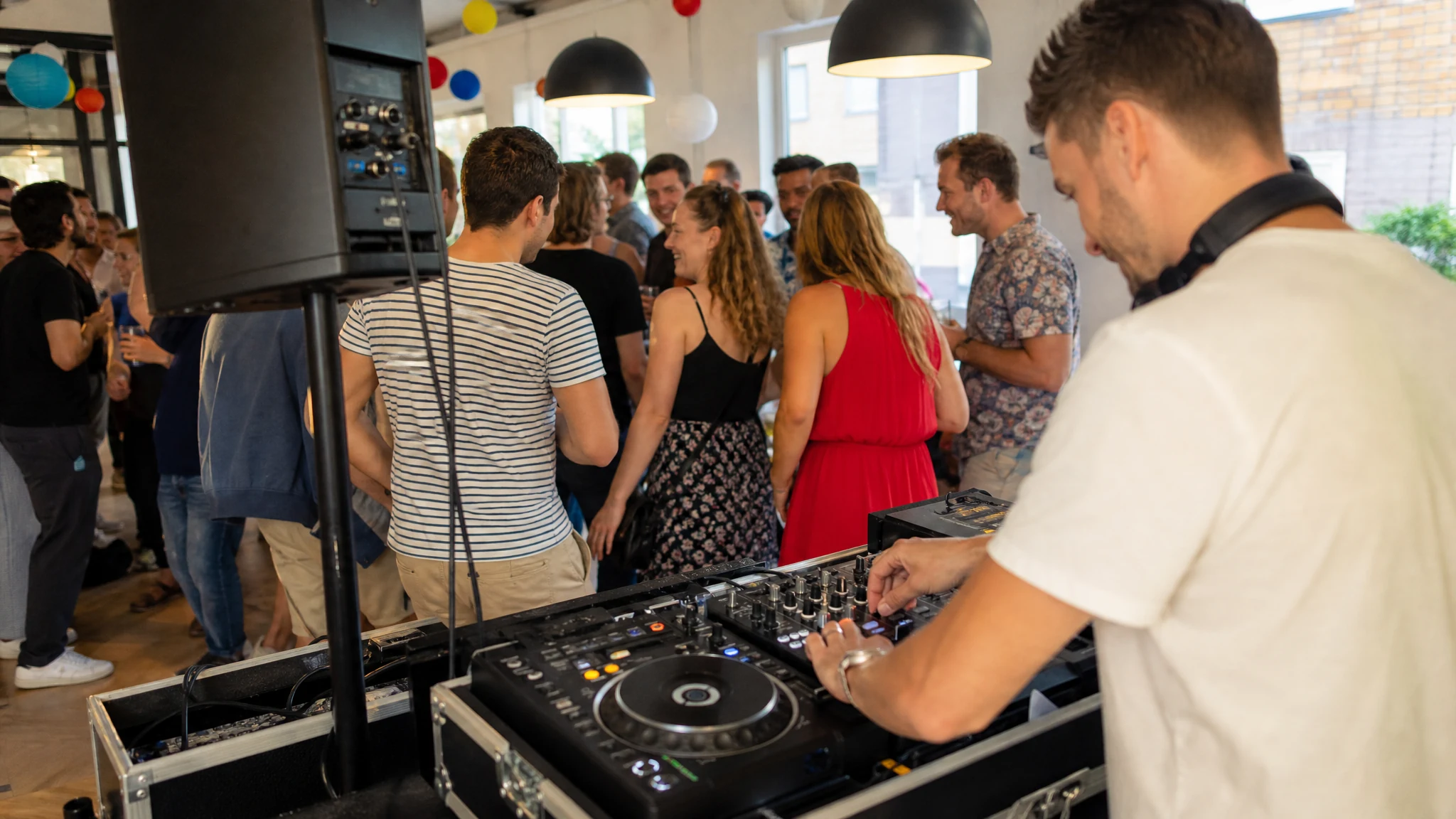 Ein DJ legt an einem Mischpult in einem hellen Raum auf, während Gäste eines Polterabends im Hintergrund plaudern