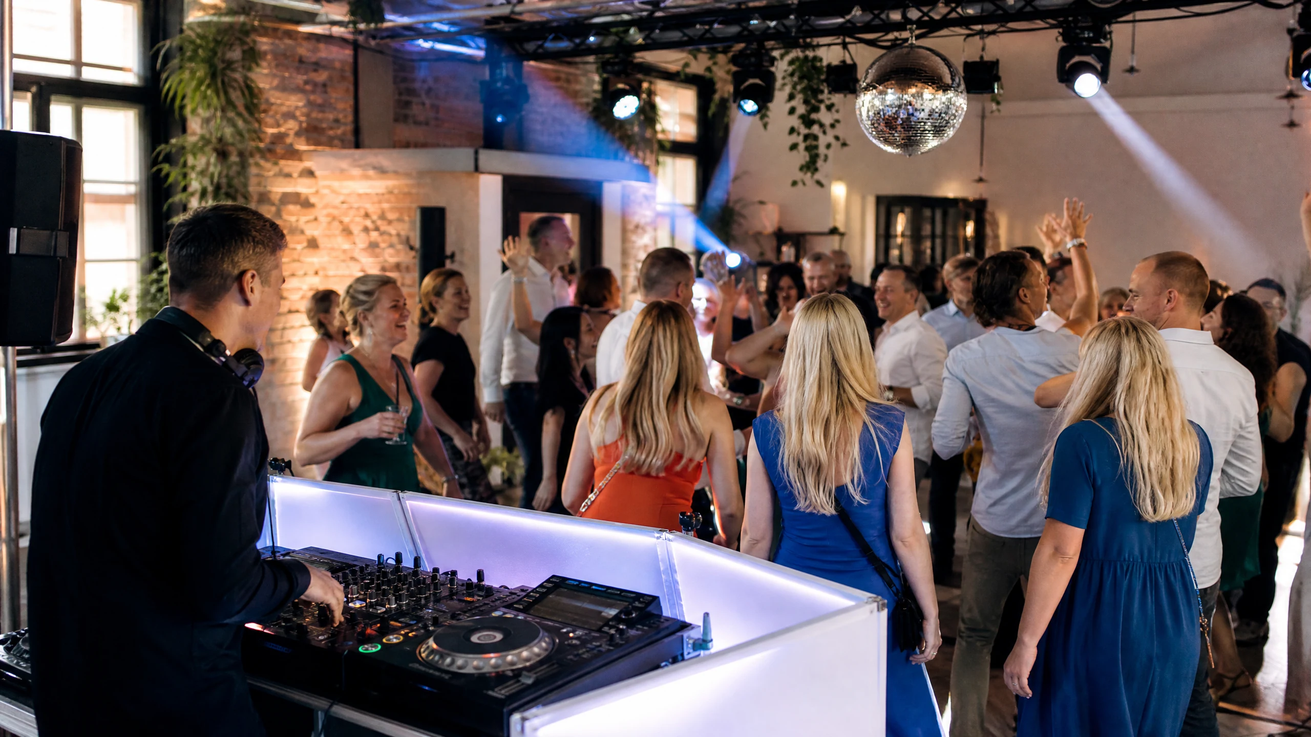 Ein DJ mit Kopfhörern legt am Pult auf, während Gäste eines Polterabends in einer Halle mit Discokugel tanzen