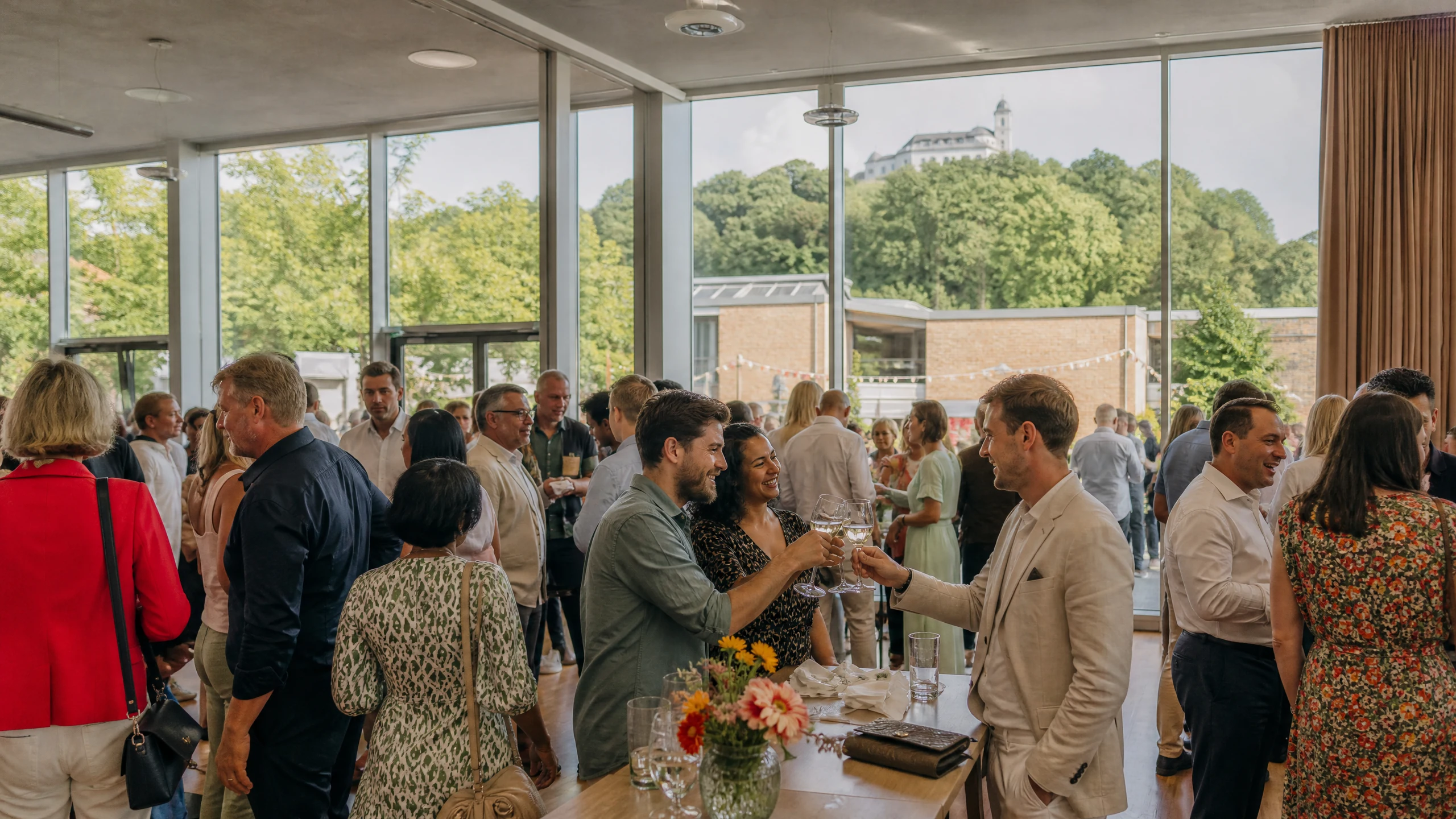 Gäste feiern in Siegburg mit Sektgläsern in einem Saal mit großen Fenstern und Blick auf bewaldeten Hügel