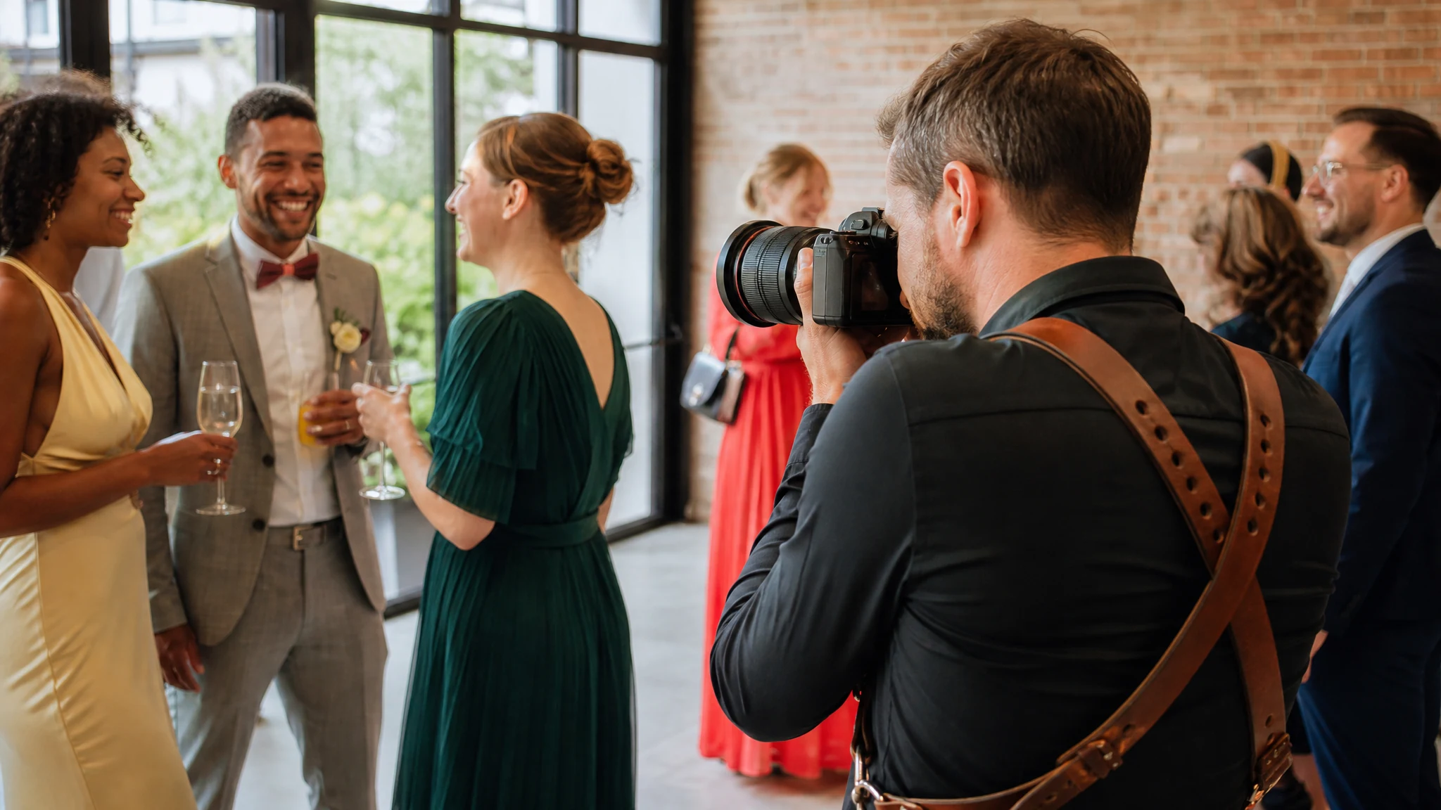 Ein Fotograf mit Kamera fotografiert lachende Gäste bei einer Hochzeit in Bonn in einer hellen Halle mit Backsteinwand