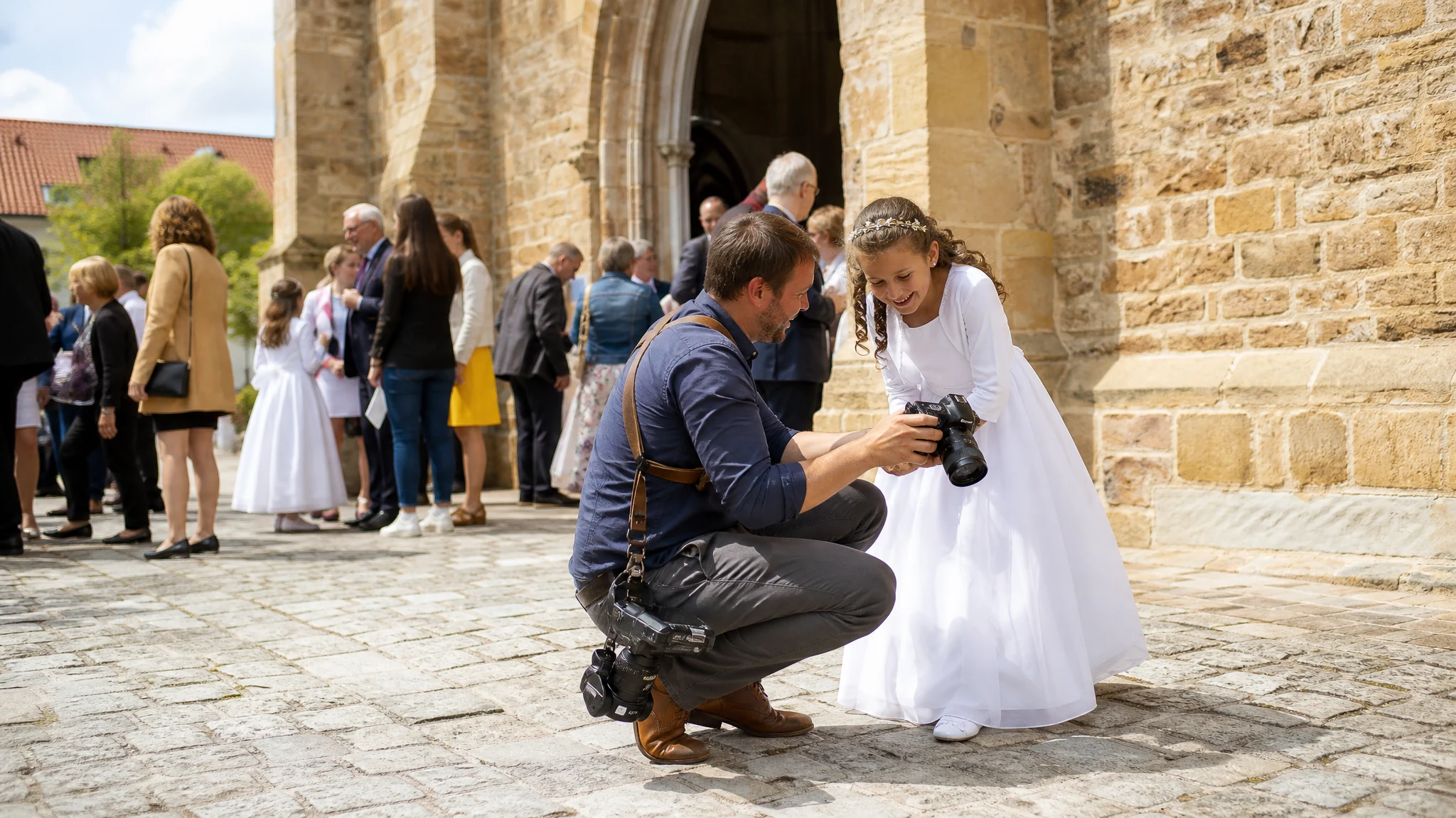 Ein Fotograf zeigt einem Mädchen im weißen Kleid vor einer Kirche die Kamera nach der Kommunion, Gäste im Hintergrund