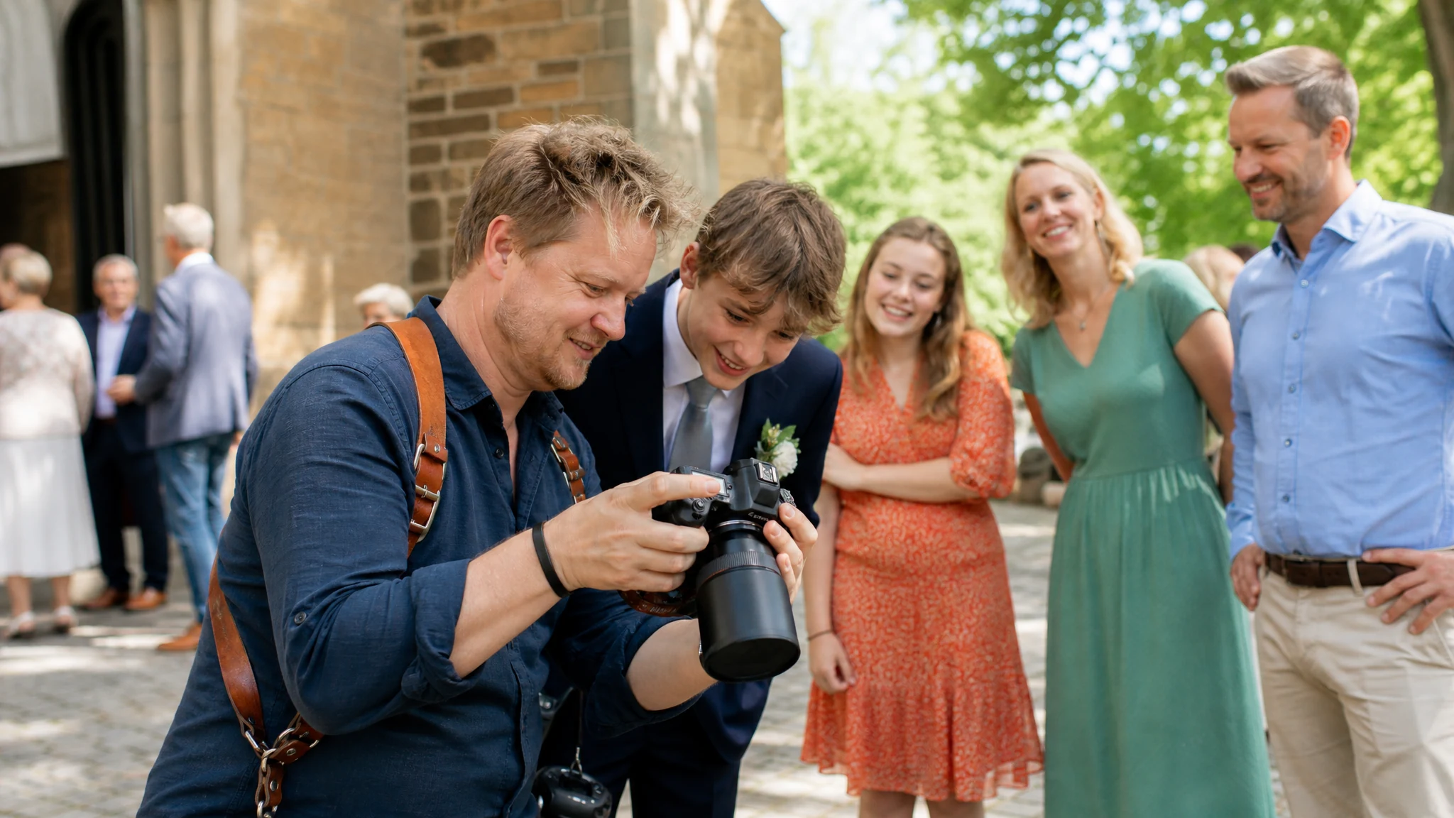 Ein Fotograf zeigt einem Jungen im Anzug die Kamera, Familie schaut lächelnd zu, bei einer Konfirmation vor der Kirche