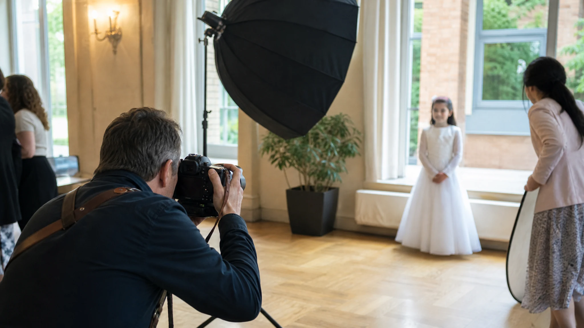 Ein Fotograf fotografiert ein Mädchen im weißen Kleid mit Softbox und Reflektor in einem hellen Saal bei einer Konfirmation