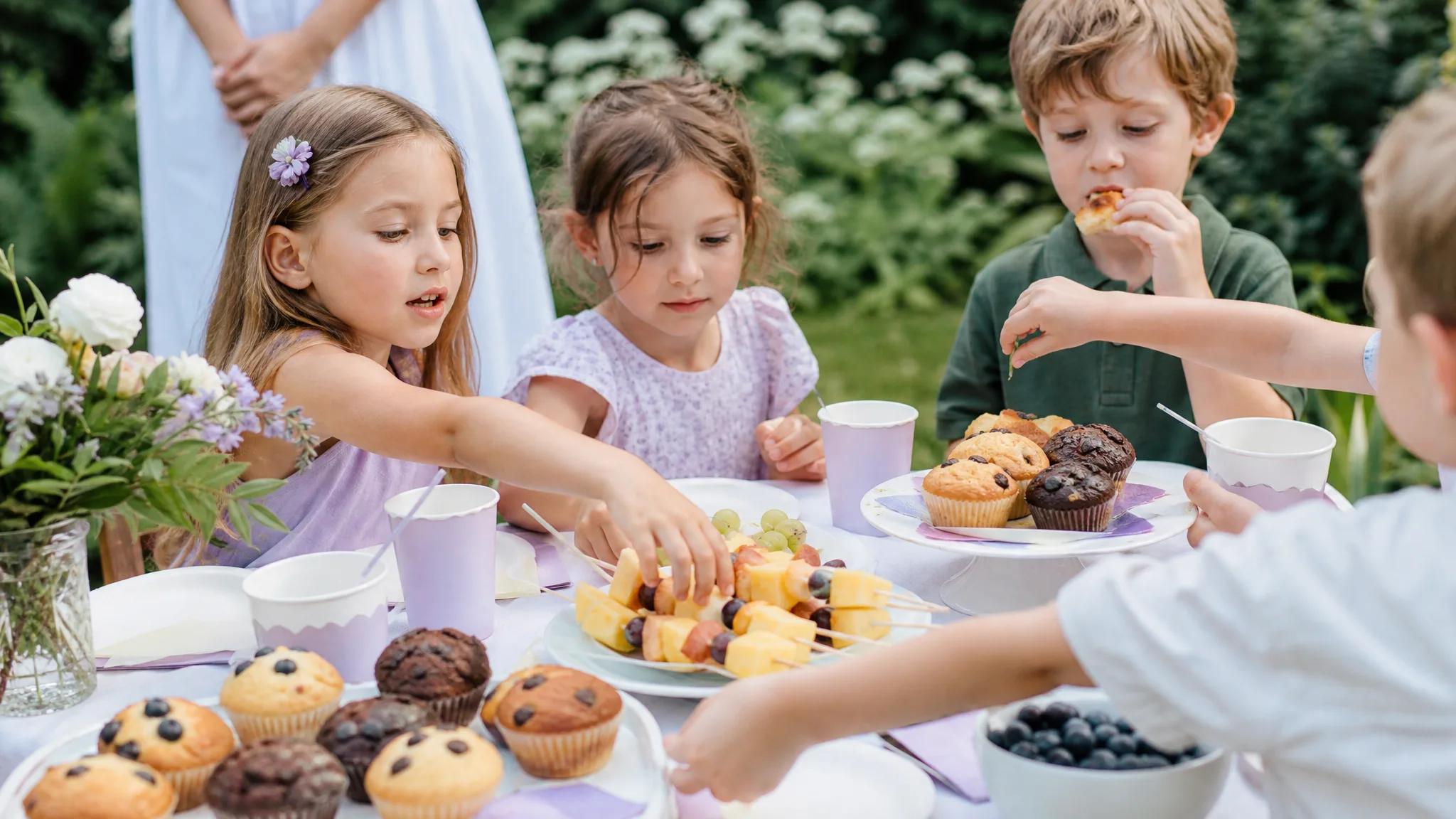 Liebevoll angerichtetes Kindergeburtstags-Buffet mit Muffins und Obstspießen, Kinderhände greifen zu