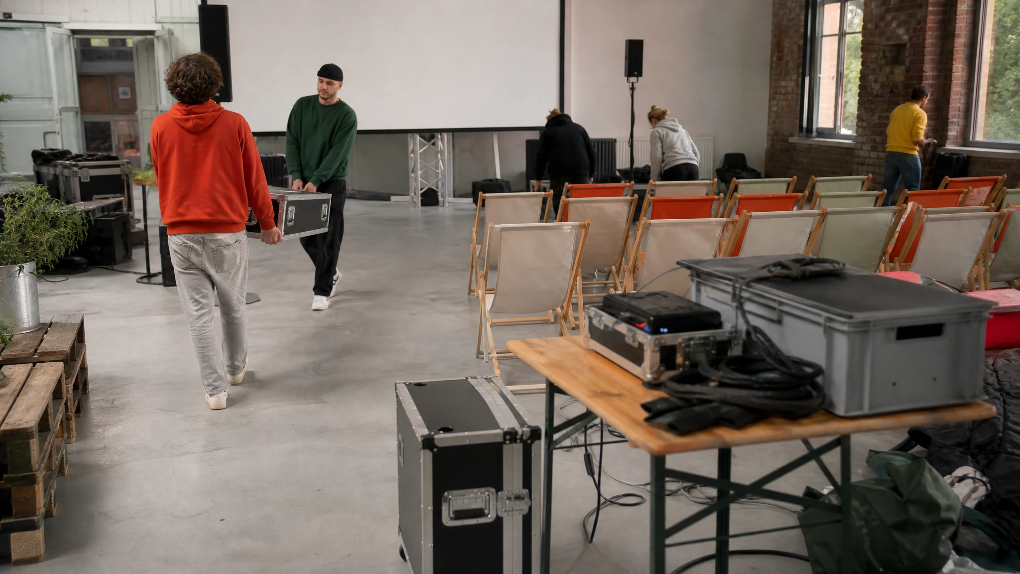 Helfer tragen Technikkoffer in eine Halle mit Liegestühlen und Leinwand, um ein Public Viewing zu veranstalten
