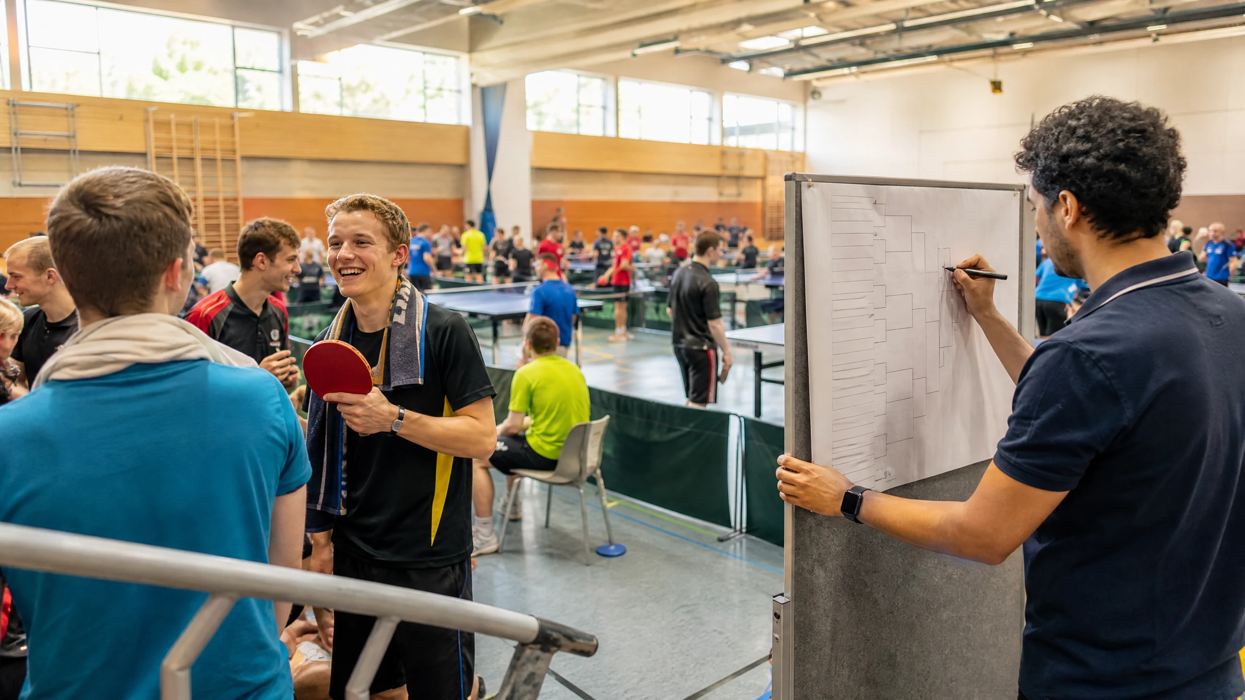 Ein Mann zeichnet in der Sporthalle einen Turnierbaum auf eine Tafel, um ein Turnier zu organisieren, ein Spieler lacht