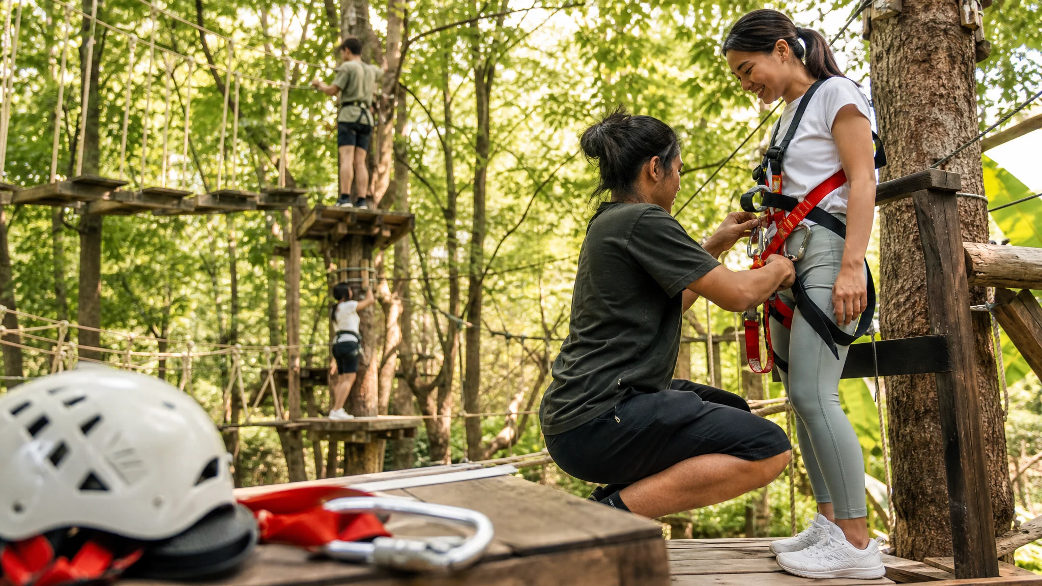 Ein Mitarbeiter legt einer lächelnden Frau im Hochseilgarten den Klettergurt an, im Vordergrund liegen Helm und Karabiner