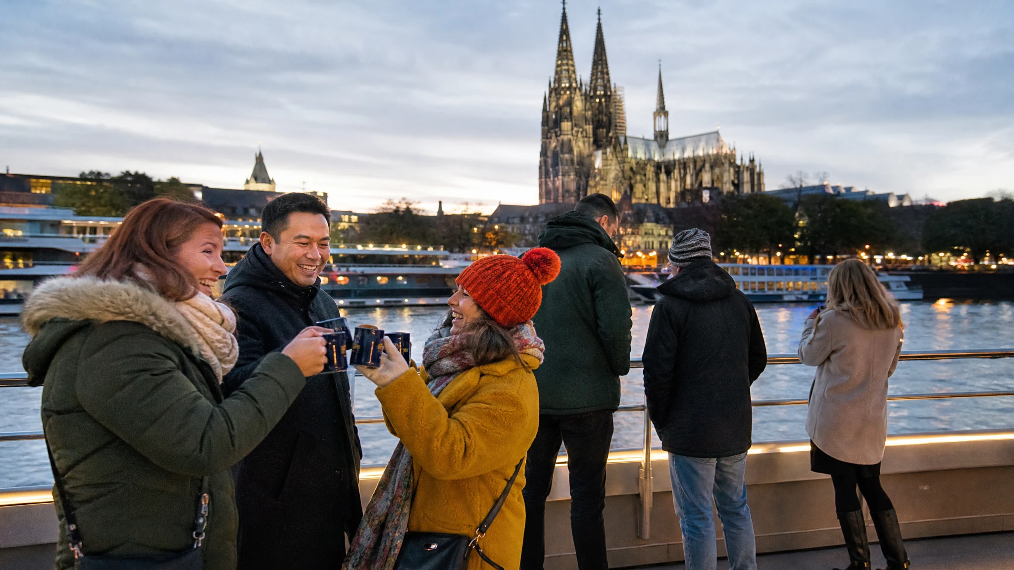 Gäste stoßen auf einem Schiff mit Tassen an, Ideen für eine Weihnachtsfeier in Köln mit Blick auf den Dom