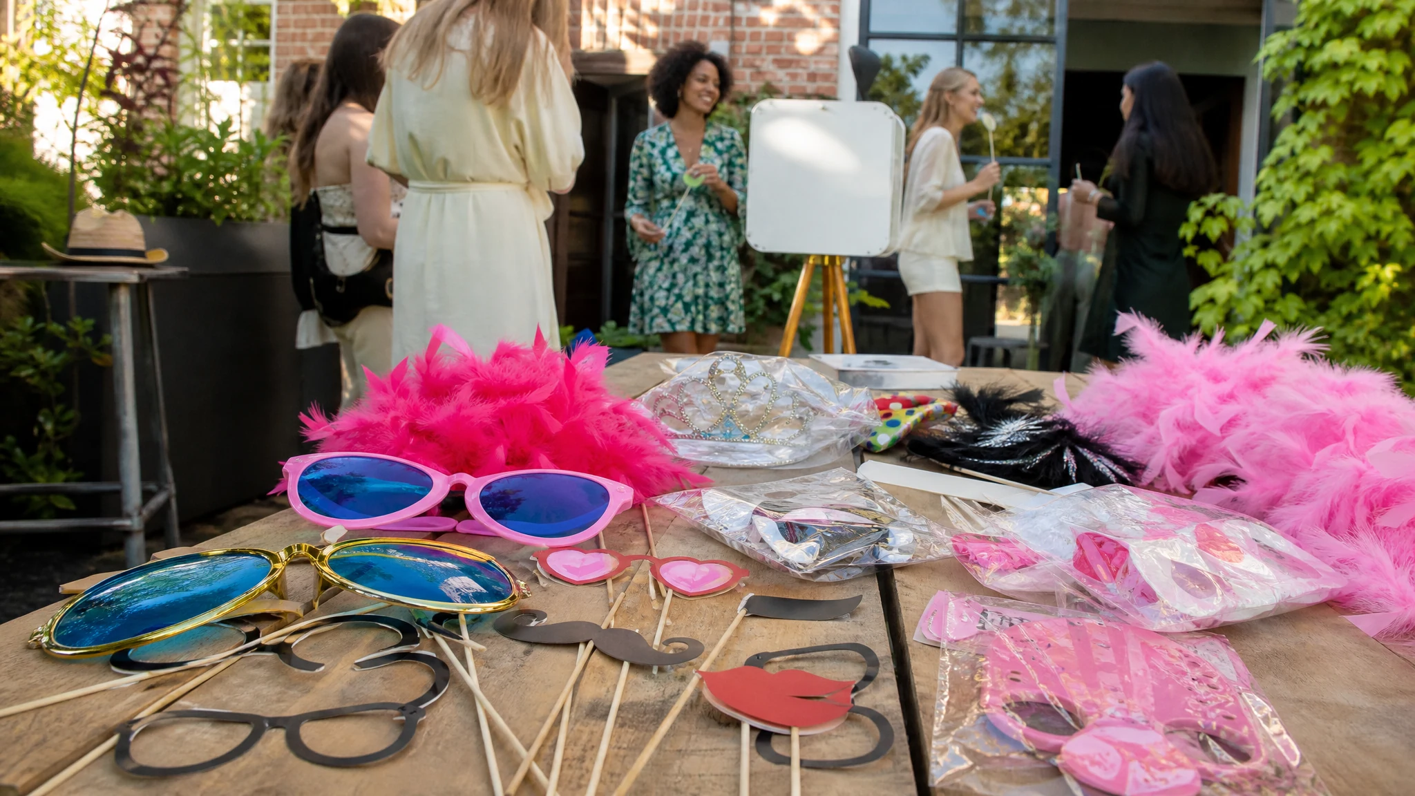 Auf einem Holztisch liegen bunte Requisiten für eine Fotobox beim JGA, während Frauen im Garten plaudern