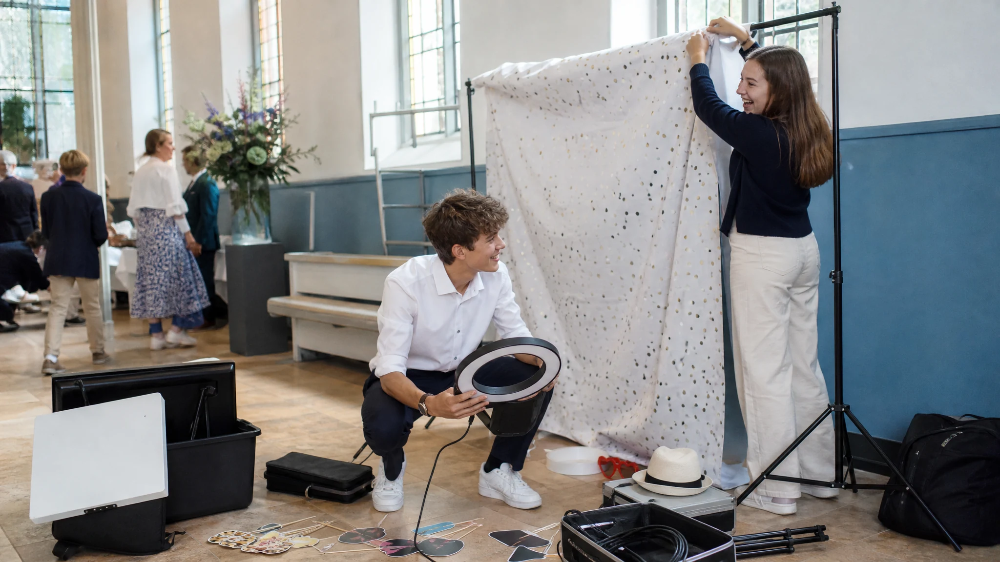 Ein Jugendlicher mit Ringlicht und eine junge Frau am Hintergrundstoff bauen eine Fotobox für eine Konfirmation im Saal auf