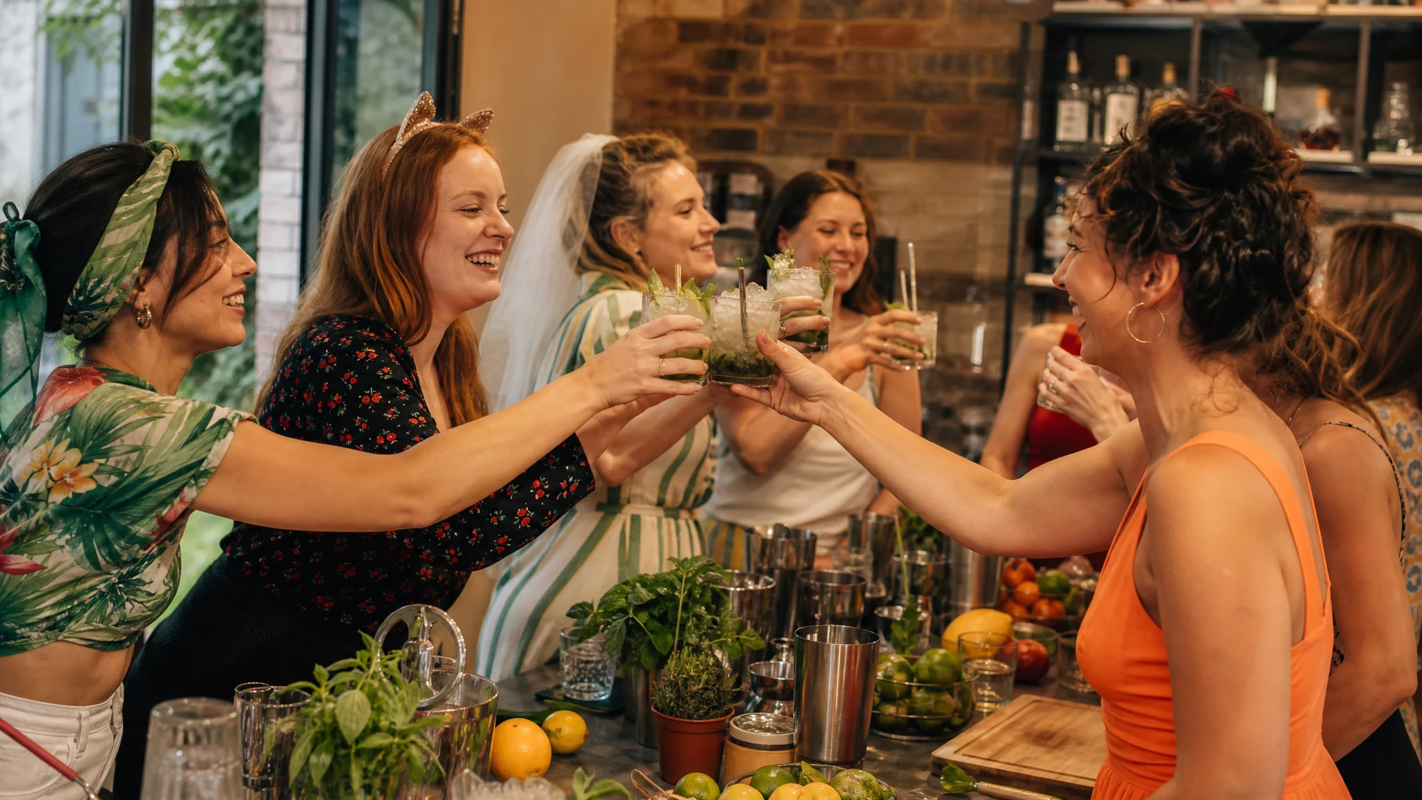 Frauen stoßen lachend mit Mojitos an einer Bar an, eine Braut mit Schleier feiert ihren JGA in einer Cocktailrunde