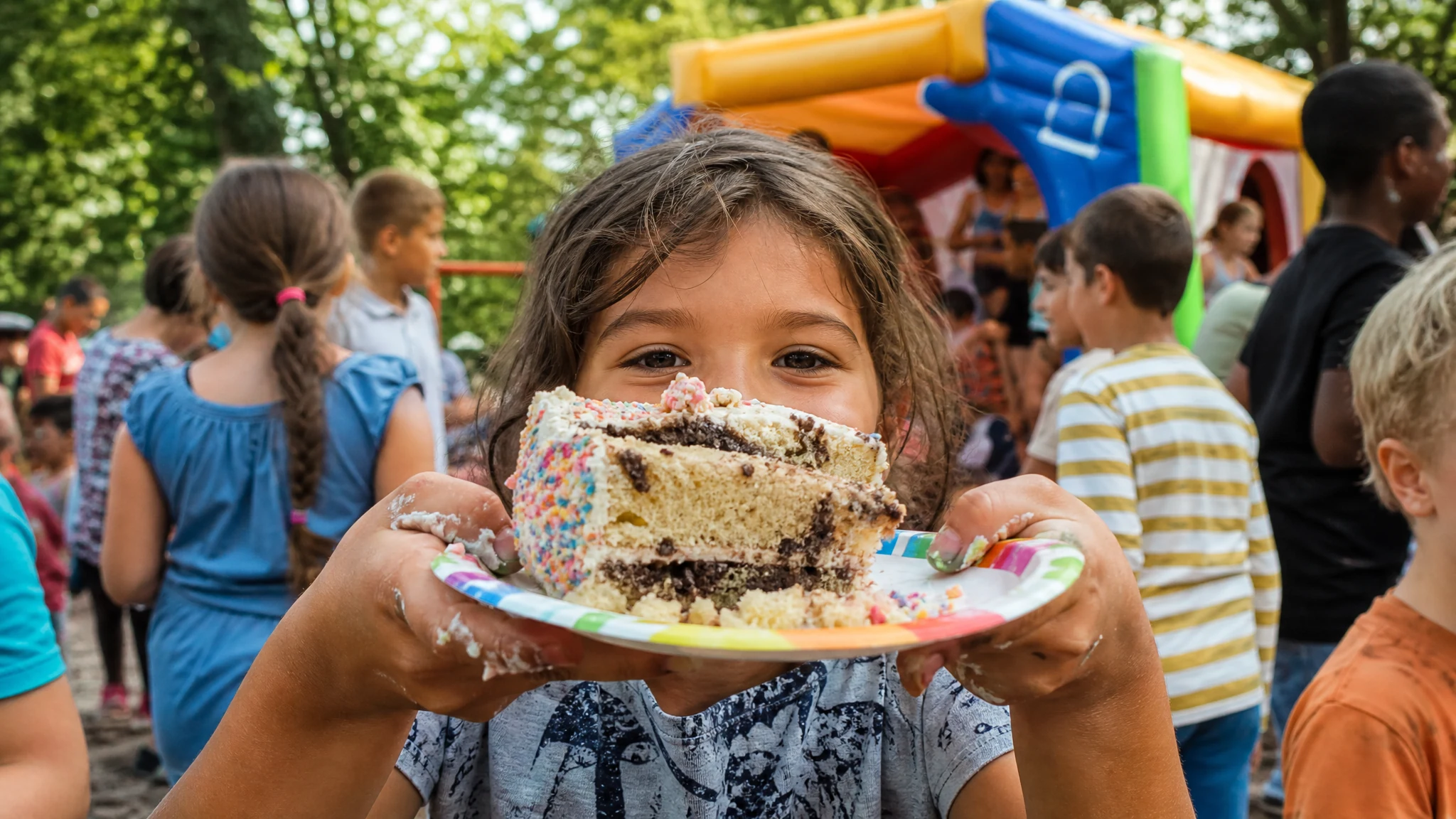 Ein Mädchen hält auf einem Kinderfest im Freien ein großes Stück Kuchen mit bunten Streuseln auf einem Pappteller hoch