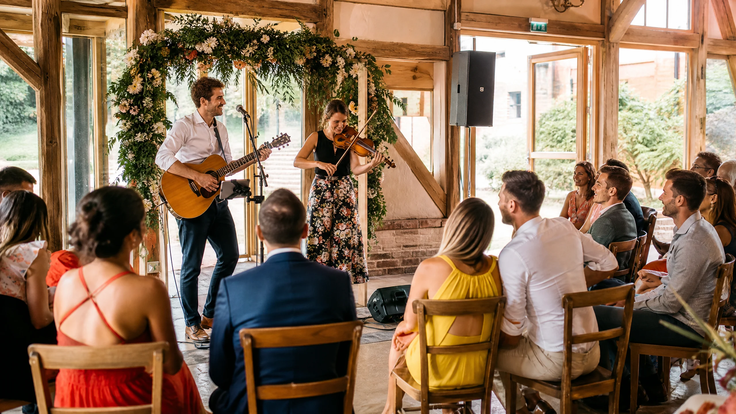 Eine Band aus der Nähe spielt mit Gitarre und Geige unter einem Blumenbogen, Gäste hören bei der Hochzeit zu