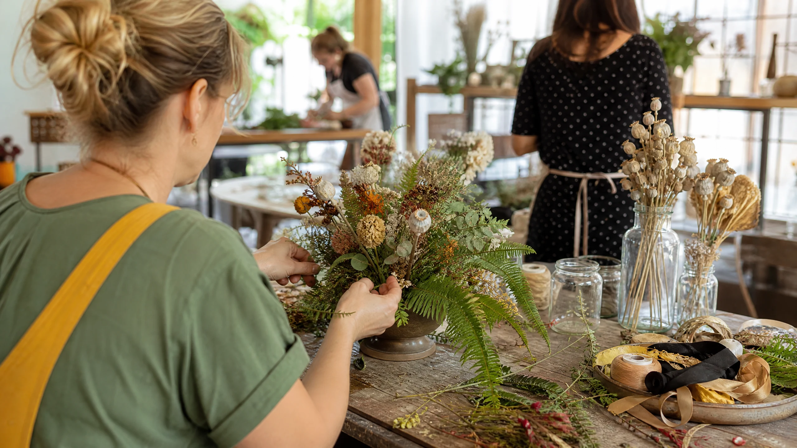 Eine Frau steckt in einer Werkstatt Farn und Trockenblumen zu einer Blumendeko für eine Hochzeit zusammen