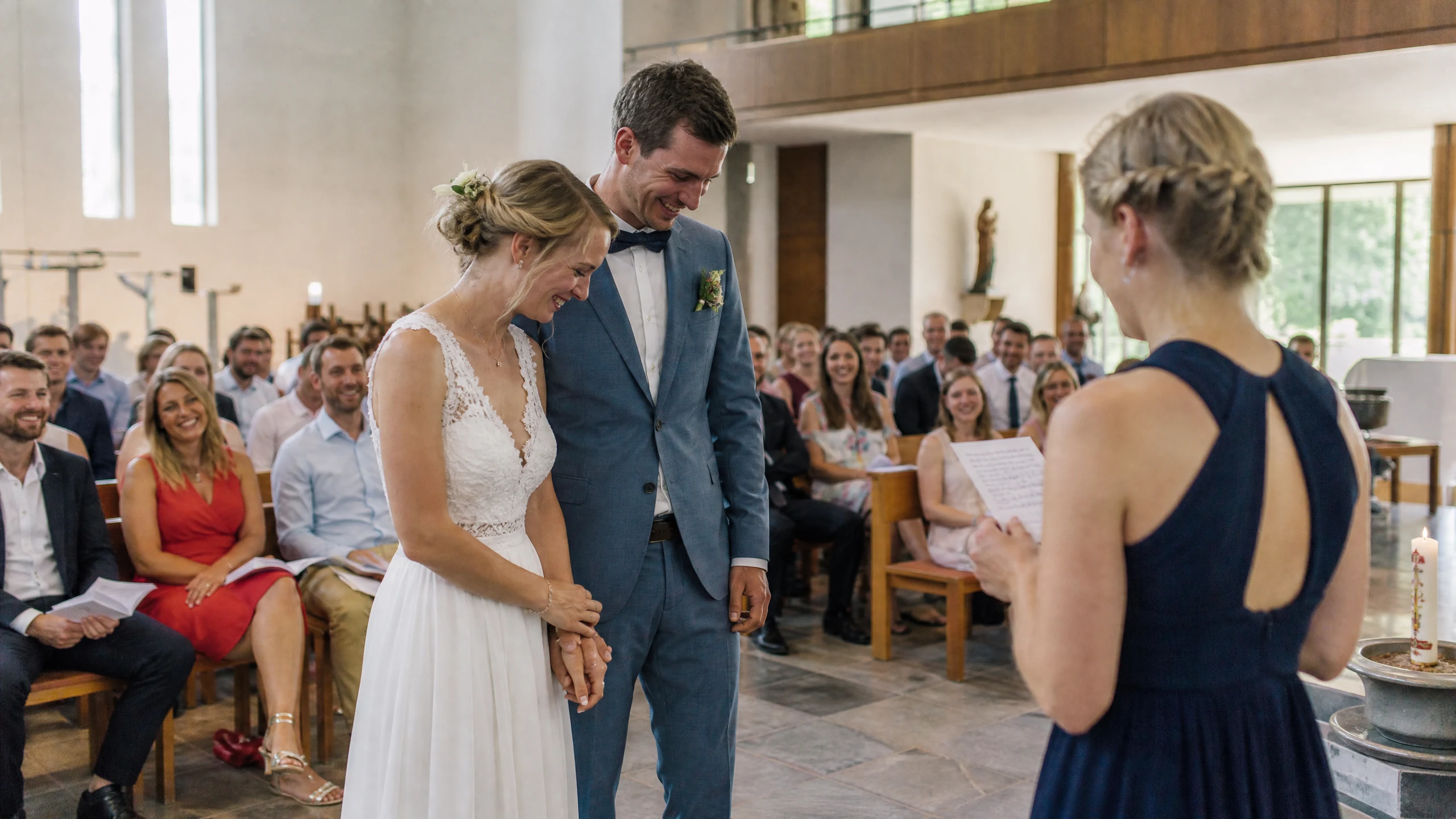 Eine Frau in Dunkelblau liest Fürbitten bei der Hochzeit in einer Kirche vor, das lächelnde Brautpaar hört zu
