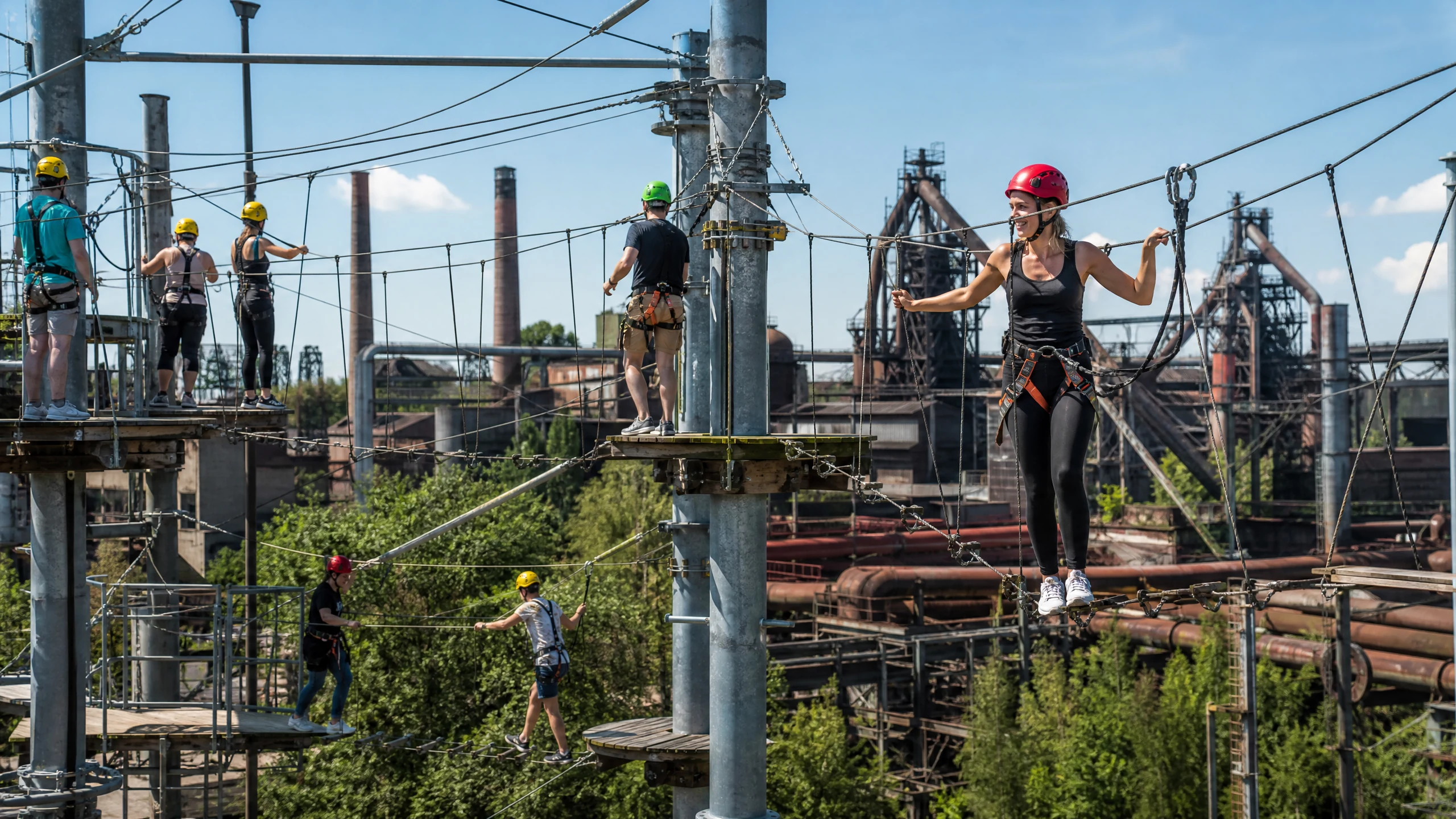 Eine lächelnde Frau mit Helm balanciert in einem Hochseilgarten in Duisburg vor alten Hochöfen, Kletterer dahinter