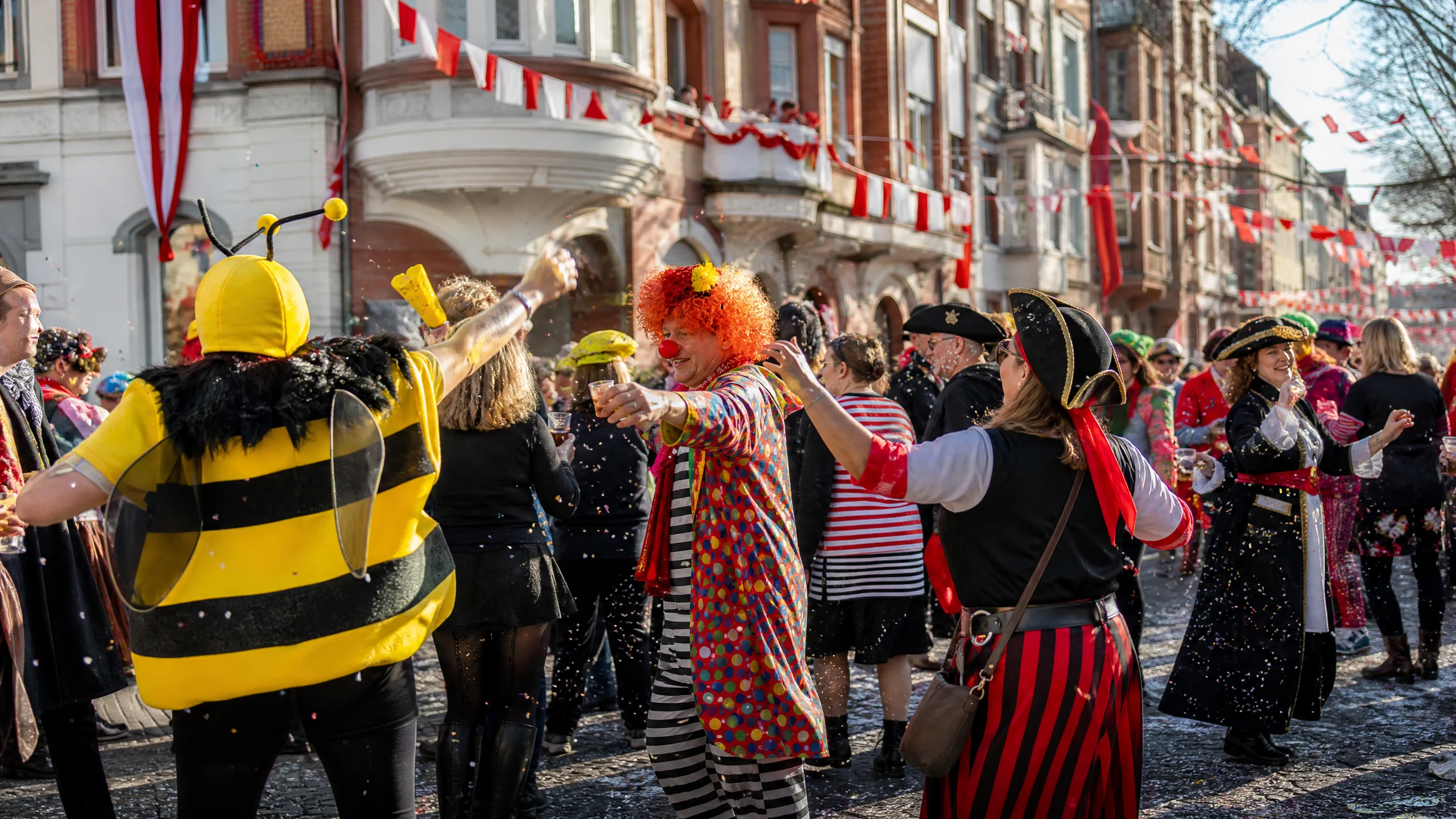 Verkleidete Feiernde als Biene, Clown und Piratin tanzen beim Karneval in Köln auf einer geschmückten Straße