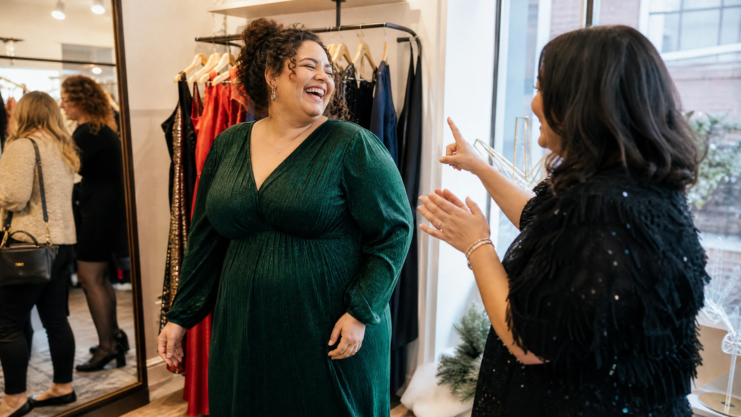 Eine lachende Frau in grünem Glitzerkleid wird in einer Boutique beraten, auf der Suche nach Kleidern für die Weihnachtsfeier